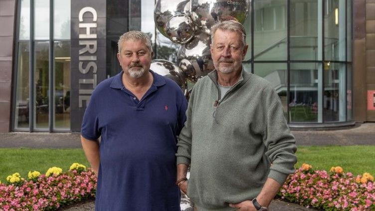 Glenn and Russell Wood are smiling as they stand outside of the main entrance to The Christie. A flower bed and a silver sculpture can be seen behind them. Glenn has grey hair and a goatee beard and is wearing a blue short-sleeved top while Russell has grey hair and a beard and is wearing a white top with a khaki green sweatshirt.