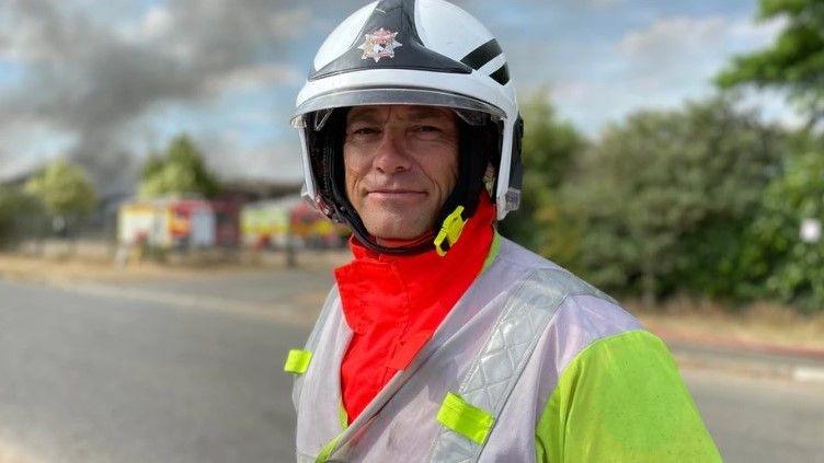 A male firefighter poses for a photograph in his helmet and uniform with fire engines in the background