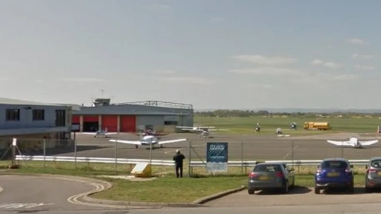 The run-way of Gloucestershire Airport with a car park in front and blue an black car parked inside.