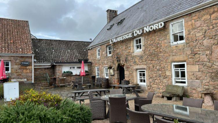 Exterior of L'Auberge Du Nord in Jersey. The stone pub has eight windows with white panels. A number of tables and chairs are outside in the empty beer garden.