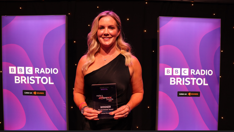 Louise Stewart with long blonde hair, holding her award and smiling at the camera. She is standing in front of BBC Radio Bristol banners.