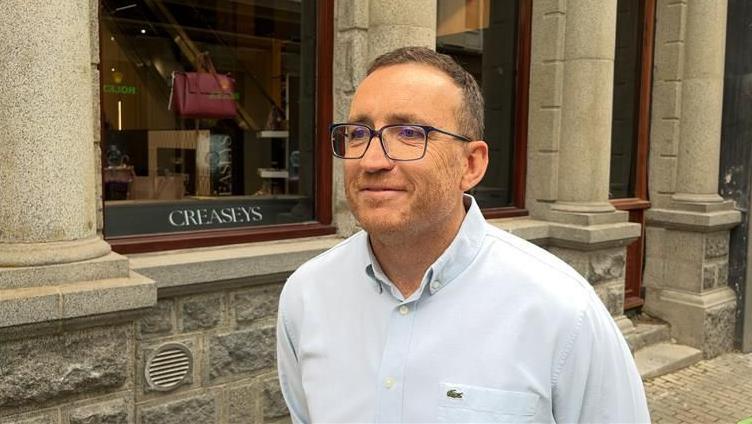 A smiling man in a blue shirt and glasses stands in front of a granite shop with pillars and a Creaseys sign in the window, along with merchandise.