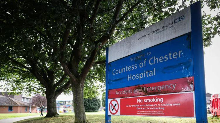 A Countess of Chester Hospital blue and red sign sits outside a hospital building with trees in the background