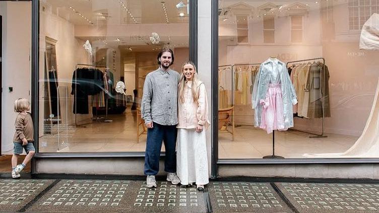 Two people are standing outside of a shopfront. The shop is white with black window and door frames. The name of the shop 'Kindread of Ireland' is printed in black letters at the top of the photo. There is also a young child standing outside the shop door to the left. On the shop window there is a large cream bow and a mannequin wearing a green top and pink bottoms. Through the shop window we can see rails of clothes inside.  