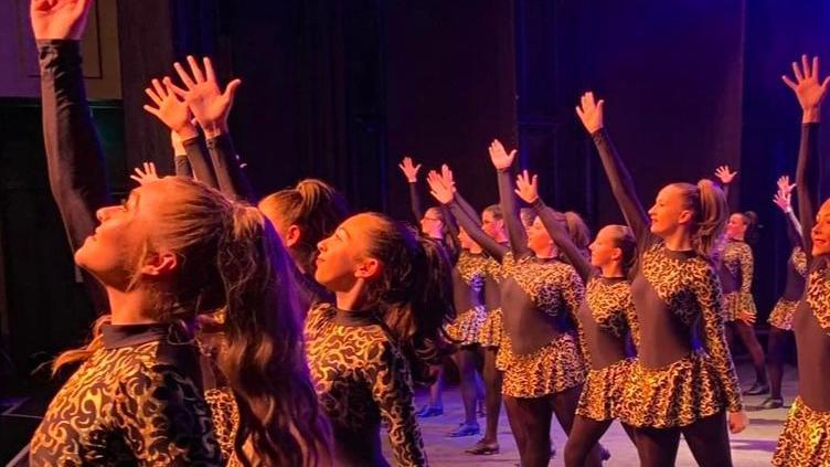 A side view of Bethan and others in cheetah-print dresses dancing with one arm extended above theme on a stage