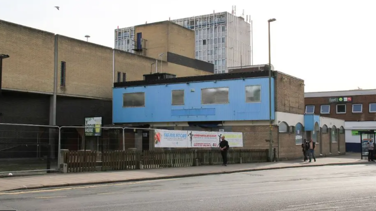 A large, 60s style brick building attached to the side of a shopping centre is seen boarded up, with metal fences separating it from the pavement.