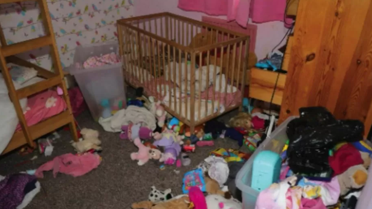 A very messy child's bedroom, with bunk beds on the left, a cot in the centre and toys and clothes strewn across the floor and a discarded bin bag on the right