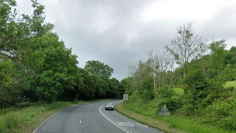 A three lane road swings through green countryside. A car drives towards us on one side of the road with the word "Slow" written on the road ahead of it. The other side is made of a lane with an overtaking lane in the middle with dotted white lines between them.