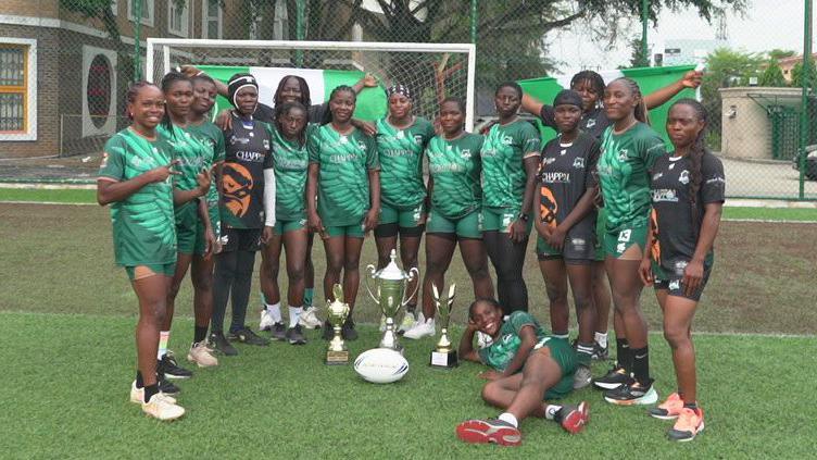 A picture showing 15 of Nigeria's Green Falcons players standing in a curved line wearing green and black kit, holding two Nigerian green and white flags up behind themselves and lining up behind three trophies and an oval rugby league ball. One of the players smiles as she lies on the ground next to the trophies and ball