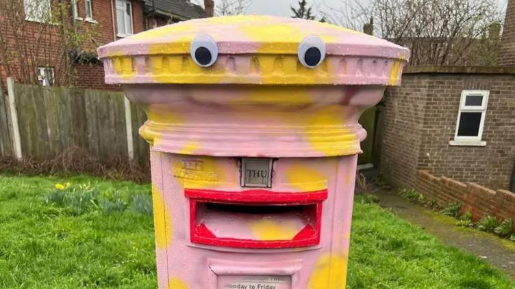 A postbox is painted pink with yellow spots. Eyes have been stuck on it and the slot for letters has been painted red to resemble lips. 