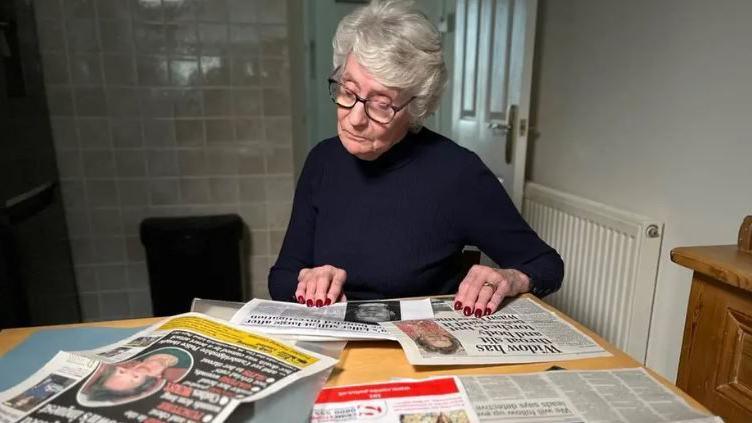 Photograph of Judy Payne, who has short grey hair  and dark-rimmed glasses, wearing a navy jumper looking at newspaper stories about her aunt's murder on the kitchen table