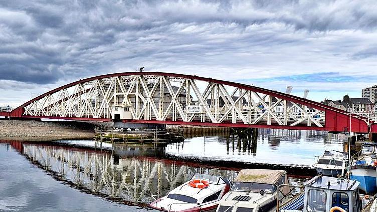 Ramsey Swing Bridge, a curved red and white structure with white x-shaped metal bars.The water is flat calm and there are a number of boats in the foreground.
