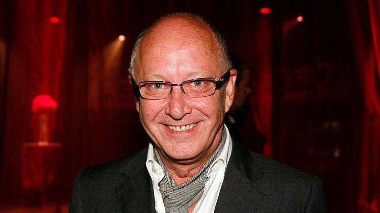 Trevor Sorbie, who has a bald head and glasses, smiles at the camera. He is wearing a black jacket, white shirt and a speckled grey cravate. There is a dark red background.