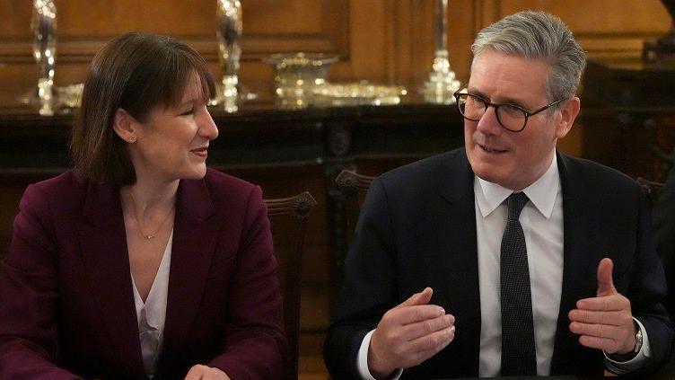 Rachel Reeves looks sideways at Keir Starmer as he is talking. Starmer is in dark business suit and tie. Reeves is in a burgundy suit. They are at an investment round table event in Downing Street.