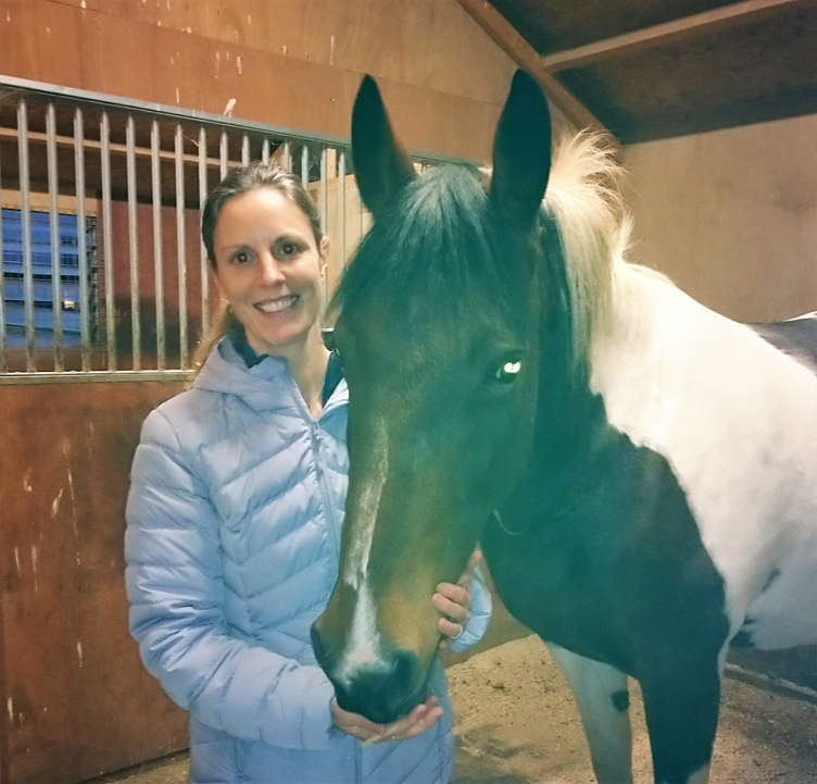 Kirstie poses happily with a horse in a stable. She wears a thick winter coat, light blue in colour. The horse has a mainly brown head, with white parts on its shoulders and legs.