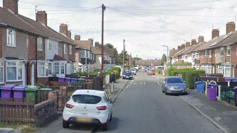 A road of terraced houses with a number of cars parked half on the pavement.