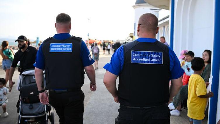 Two men walking wearing black uniform and a blue badge that reads community safety accredited officer.