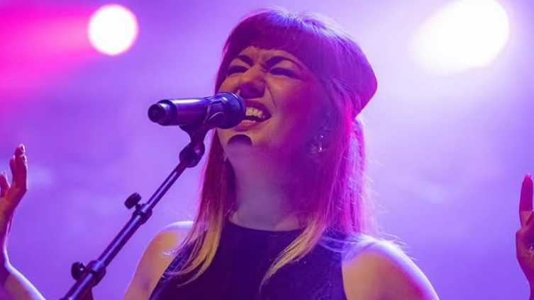 Hayley Sanderson sings into a microphone. She looks passionate and has her arms open and her eyes closed. The background is bathed in purple lighting.