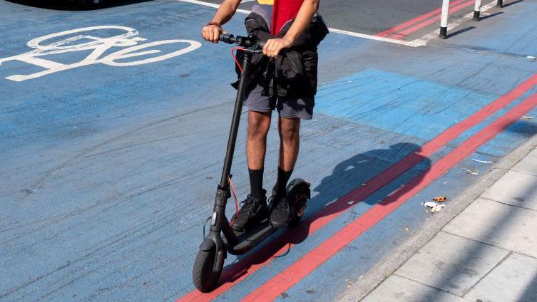 Man shown from neck down riding an e-scooter in a cycle lane on a London road.