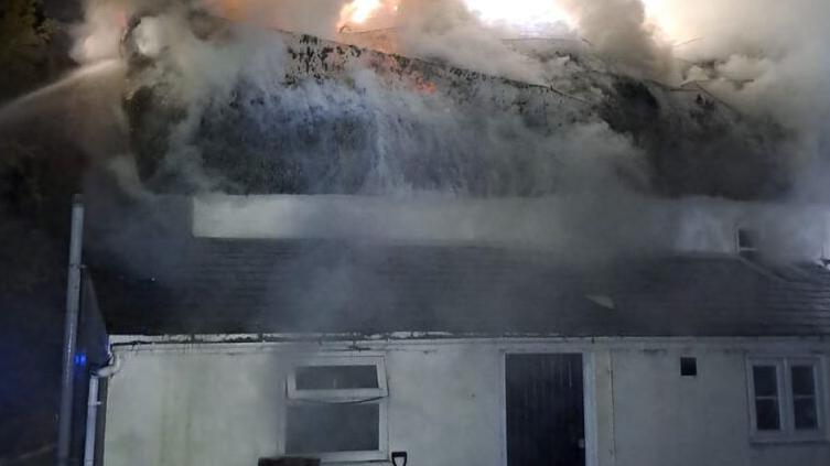 White cottage with thatched roof on fire and smoke pouring out.