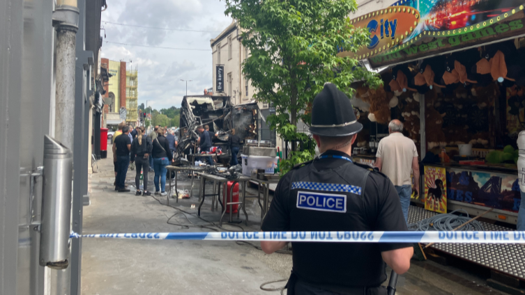 A policeman looks down the street in Hereford towards the burnt out refreshment stall which has been destroyed in the fire. It is near to a fairground ride and some people are gathered by it. There are buildings on either side of the street.