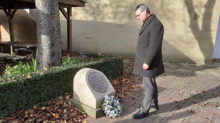 A man, Councillor Mark Ieronimo, wearing a smart grey coat and trousers bows his head towards a memorial with a wreath on. The granite memorial is beneath a tree and autumn leaves surround it.