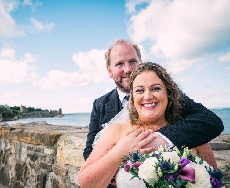Lori and Jacqui. He is in a light grey formal suit and has his arms around Jacqui who is in a wedding dress. She is smiling and holding flowers. They are pictured next to a stone harbour wall next to the North Sea.