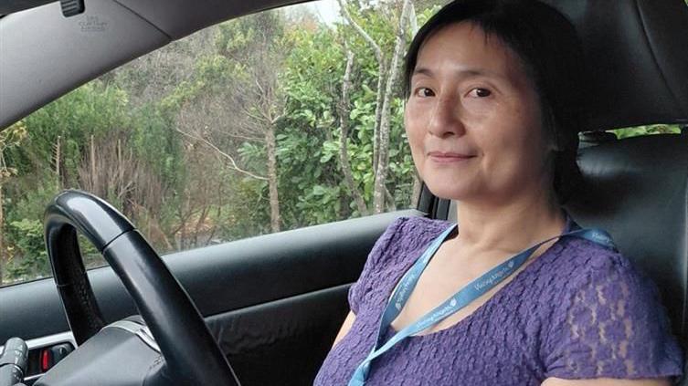 Poling Choi is wearing a purple top and blue lanyard. She is sitting in the driving seat of her car. The car is parked near countryside and there are trees in the background.