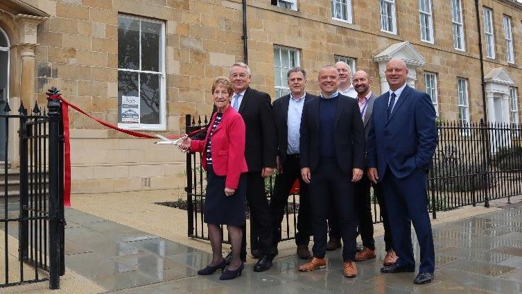 Mayor of North Tyneside Dame Norma Redfearn in a black skirt, red blazer and black and white blouse, cutting a red ribbon outside a stone building. Standing next to her right is Tynemouth MP Sir Alan Campbell, architect Tim Brook, Compass Developments Director Stephen O’Neill, North Tyneside Council Project Manager Fergus Mitchell, North Tyneside Council Housing Growth.