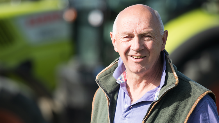 Farmer Tim Scott. He is bald with short grey hair at the sides. He is sun-tanned and is wearing a green sleeveless warmer and a blue top. The background, which shows farm machinery, is out of focus.