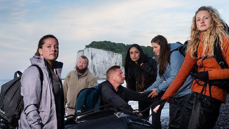 Six people - four women and two men - around a small boat on a Dover beach, for a promotional photo for Channel 4's Go Back To Where You Came From 