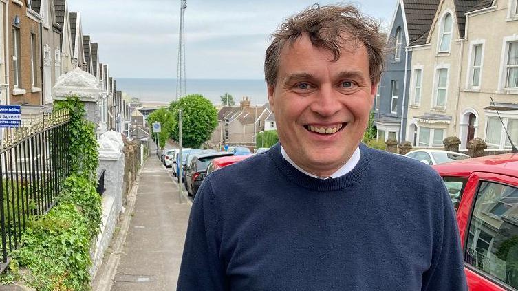 56-year-old Peter May is looking directly at the camera and smiling. You can see his teeth.
He has wavy hair and a round face.
Peter May is standing on a street that is on a hill with a view of the sea behind.