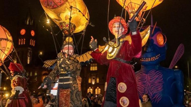 Halloween parade with large puppets in colourful costumes with people in the background looking on.
