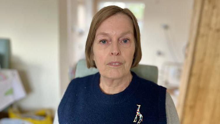 Woman with bobbed hair, wearing a blue and grey jumper. She is standing in a kitchen.