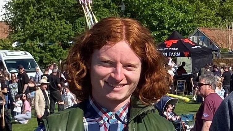George Gilbert is wearing a navy shirt with large white and red checks and a dark green bodywarmer. Charlie is wearing a dark grey jumper. Behind the brothers - who are both smiling - are lots of people gathered on the village green during an event.