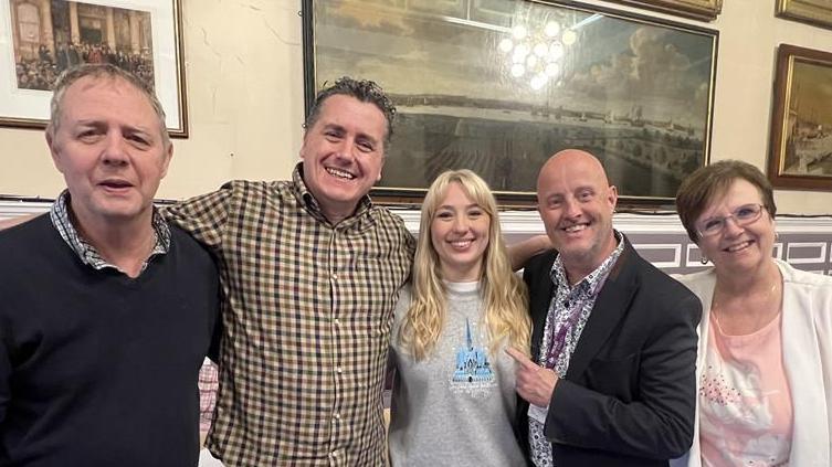 Ms Roper stands in the middle of three men and another woman who sat in her audition. A man on the left has short grey hair, blue jumper and shirt on. A man to his right wars a chequered yellow and green shirt. Ms Roper stands in the middle wearing a grey jumper. A man to her right is bald and wears a black jacket with a floral shirt underneath and a woman stands to his right with short red hair wearing a white jacket and pink top underneath. 