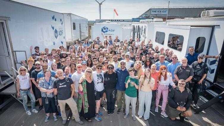 A large group of people in colourful clothing look towards the camera. There are white TV production motorhome vehicles on either side of them and behind them. A low rise building can be seen in the background.