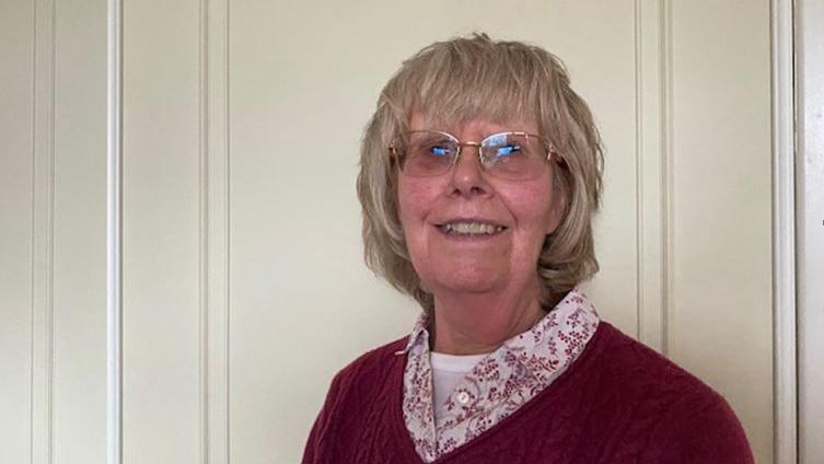 Pat Costain, a woman with short grey hair, wears glasses and a pink jumper in front of a white door