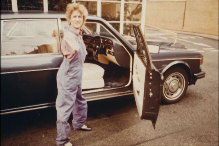 Alison Lapper, wearing a red t-shirt and blue dungarees, stands in front of a black car. The driver's seat of the car is open. 