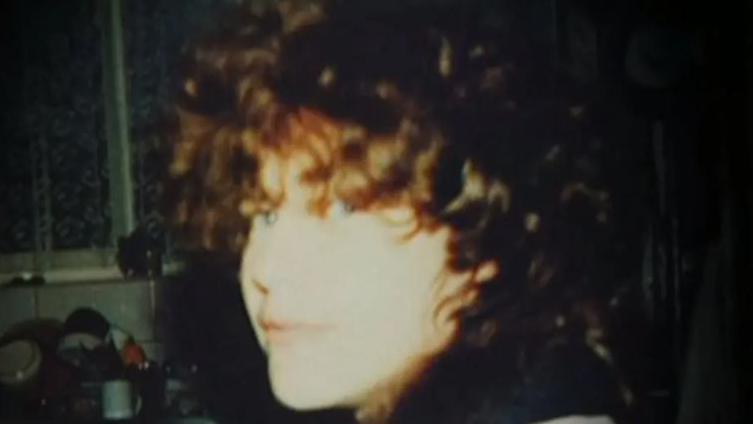 It is a grainy image of a woman, likely pictured in the late 80s or early 90s. She has curly brown hair and is slightly smiling at the camera, her eyes peeking out under a curly fringe. She is in a kitchen and washing up can be seen on the side.