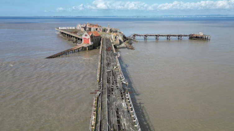 Birnbeck Pier from the air as a raised front view. The sky in the background is blue and bright with some white clouds in the sky. The tid is in and the pier is surrounded by water.