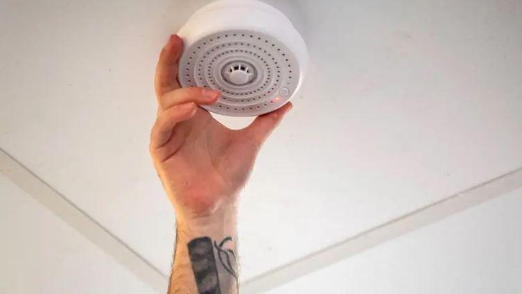 A man installing a vape detector in a school building, with his arm reaching up to the ceiling to connect the device. 