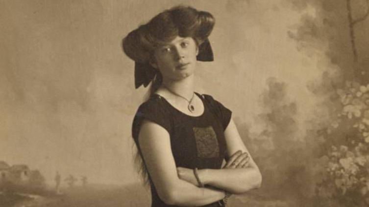 Portrait photograph of Irene Steer pictured bare foot in a swimming costume. She has her arms crossed by her waist and looks directly at the camera. She has brown hair and wears a large pendant necklace. 