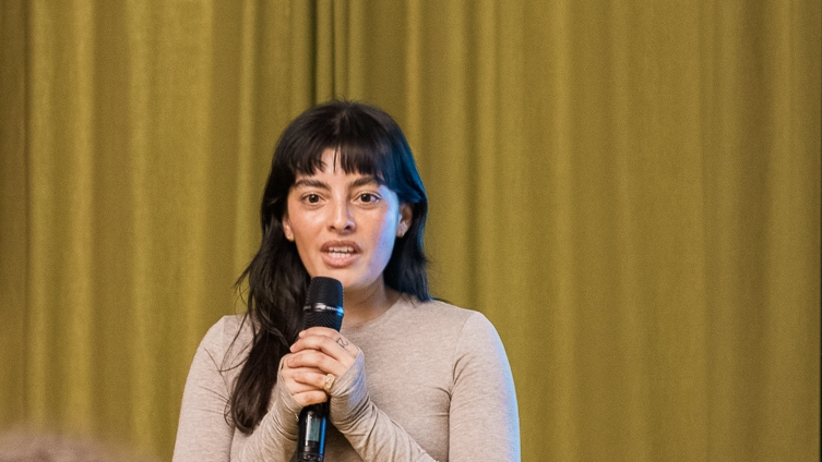 Kalpana Arias, a woman with long dark hair and a fringe, speaking into a microphone with both hands in front of a gold curtain backdrop