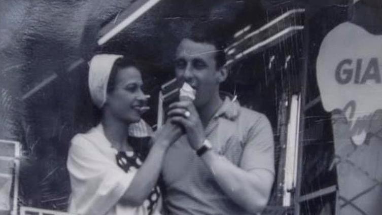 A young woman with long dark hair wearing a white hairband and spotty dress is holding an ice cream, that the young man next to her his licking. He is wearing a t-shirt with a collar. Next to them is a giant wooden ice cream, as tall as they are