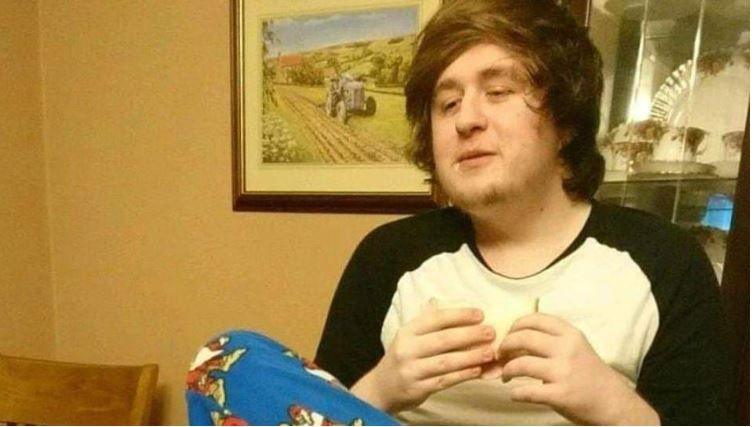 Alexander McCartney sitting in a dining room, he is wearing pyjamas. He has long dark, fuzzy hair. There is a picture of a countryside scene with a tractor in the background. He is eating a sandwhich.