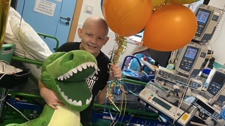 Harley, who is wearing a black T-shirt, holds a large teddy dinosaur and balloons. He has a shaved head due to his chemotherapy and is standing in front of hospital equipment and technology. He is smiling.