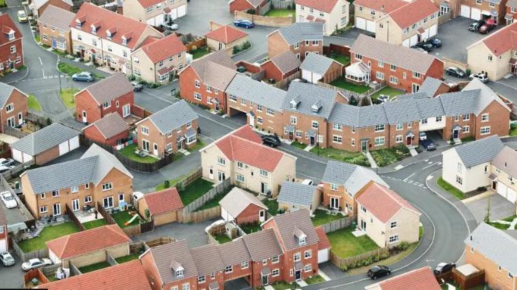 A birds eye view of a housing estate, with red roofed houses with neat gardens dotted around a grey road