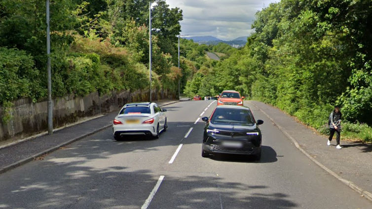 Three cars drive along Clune Brae while a woman walks on the pavement