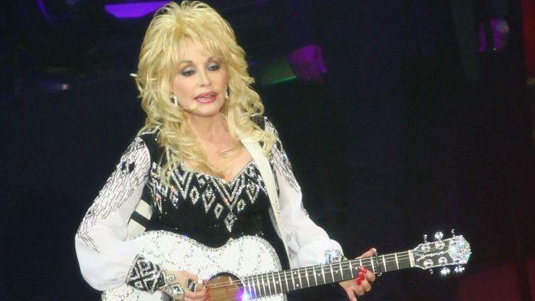 Holding a glittering white guitar, Dolly Parton stands on stage wearing a black dress with wide sleeves.